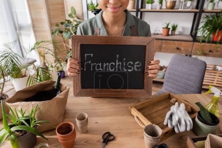 partial view of african american florist holding board with franchise lettering near flowerpots and gardening tools on table