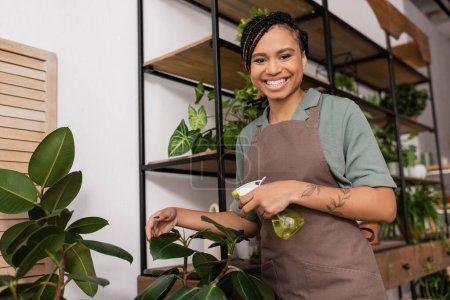 happy and stylish african american florist looking at camera while standing with spray bottle near plants in flower shop