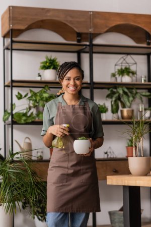 cheerful african american florist in apron holding spray bottle and potted plant while looking at camera in flower shop