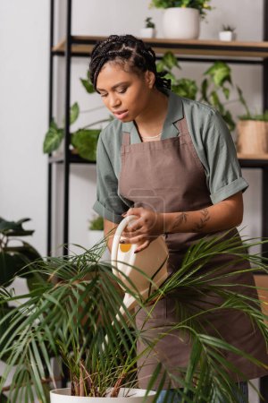 junge tätowierte Afroamerikanerin in Schürze gießt tropische Pflanze in Blumenladen