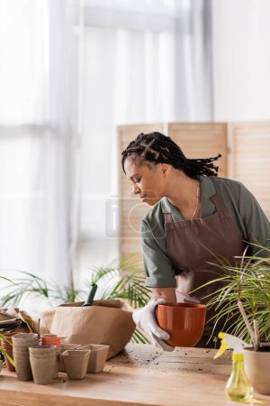 élégant fleuriste afro-américain tenant pot de fleurs et regardant dans un sac en papier avec de la terre tout en travaillant dans le magasin