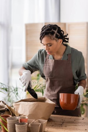 Photo for Young african american florist holding flowerpot and garden scoop near paper bag with soil - Royalty Free Image