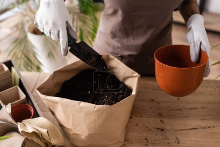 Photo for Partial view of african american woman holding garden scoop and flowerpot near paper bag with soil - Royalty Free Image