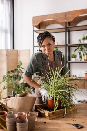 sonriente florista afroamericano mirando a la cámara mientras trabaja con plantas en la tienda de flores