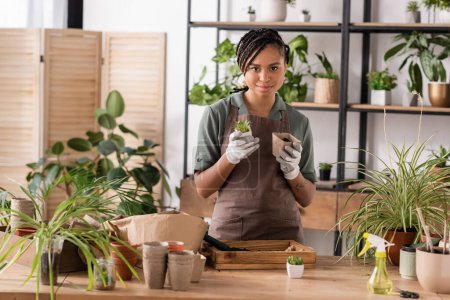 stylish african american florist in work gloves looking at camera near plants and flowerpots on table