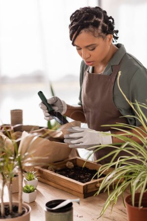 afrikanisch-amerikanischer Florist mit trendiger Frisur, der Gartenschaufel und Blumentopf hält, während er in der Nähe von Pflanzen im Geschäft arbeitet