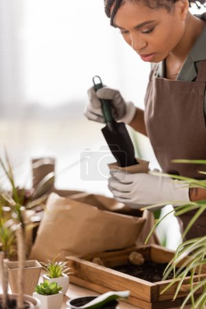 african american florist holding garden scoop and flowerpot near wooden box with soil and blurred plants