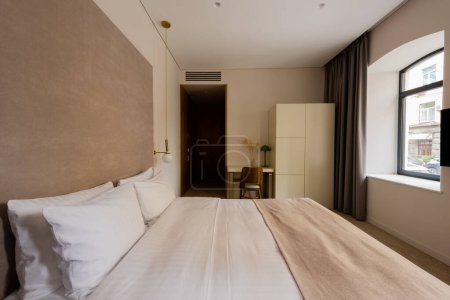 Téléchargez les photos : Comfortable bed with white bedding near desk and chair in modern hotel bedroom - en image libre de droit
