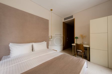 Téléchargez les photos : Comfortable bed with white bedding near desk and wooden chair in modern hotel bedroom - en image libre de droit
