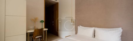 comfortable bed with white bedding near desk and chair in modern hotel room, banner 