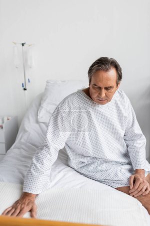 Upset senior patient in gown sitting on hospital bed
