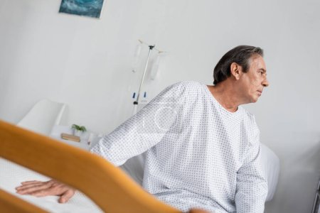 Foto de Side view of grey haired patient sitting on bed in clinic - Imagen libre de derechos
