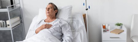 Foto de Elderly man in patient gown lying near intravenous therapy and pills in clinic, banner - Imagen libre de derechos