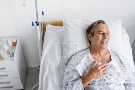 Photo for High angle view of smiling elderly man in patient gown lying on bed in hospital ward - Royalty Free Image