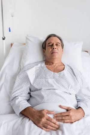 kranke grauhaarige Patientin im Kittel blickt in die Kamera, während sie in der Klinik auf dem Bett liegt