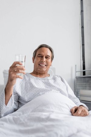 Lächelnder Mann hält ein Glas Wasser in der Hand und blickt auf dem Krankenhausbett in die Kamera