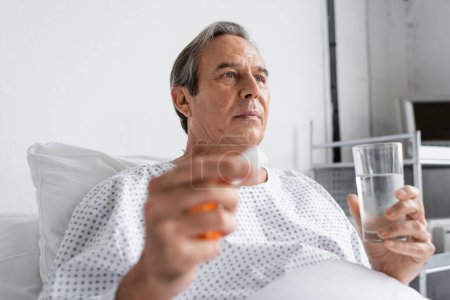 Téléchargez les photos : Senior man in gown holding pills and water in hospital ward - en image libre de droit