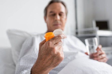 Photo for Blurred senior patient holding pills and glass of water in hospital ward - Royalty Free Image