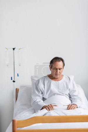 Photo for Upset elderly patient in gown sitting on bed near intravenous therapy in clinic - Royalty Free Image