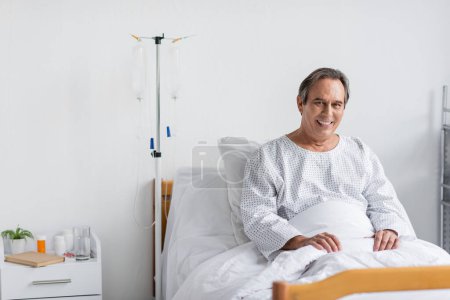 Cheerful elderly patient looking at camera while sitting near intravenous therapy in clinic