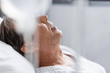 Kranker älterer Patient liegt neben verschwommener intravenöser Therapie im Krankenhaus