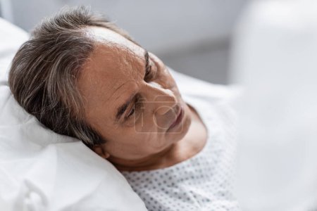 Sick grey haired man looking away while lying on bed in clinic