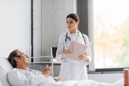 Grauhaarige Patientin im Gespräch mit Arzt mit Papiermappe in Klinik