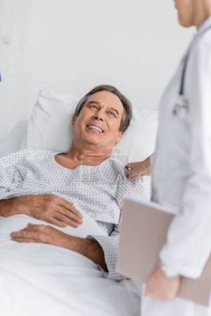 Verschwommener Arzt mit Papiermappe beruhigt ältere Patientin auf Krankenhausstation
