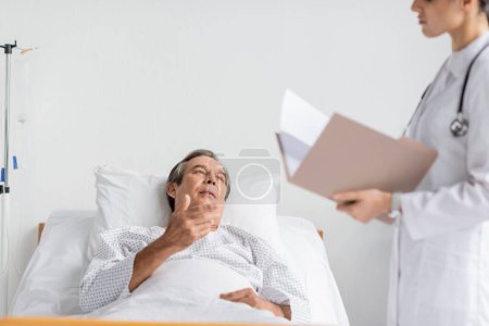 Photo for Elderly patient talking to blurred doctor with paper folder in hospital ward - Royalty Free Image