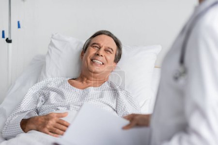 Photo for Cheerful patient in gown looking at blurred doctor with paper folder in hospital ward - Royalty Free Image