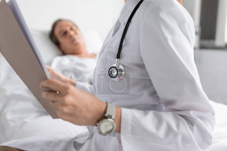 Téléchargez les photos : Doctor in white coat holding paper folder near blurred patient in hospital ward - en image libre de droit