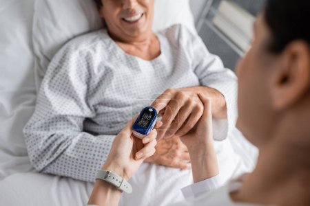 Blurred doctor wearing pulse oximeter on finger of smiling patient in hospital ward 
