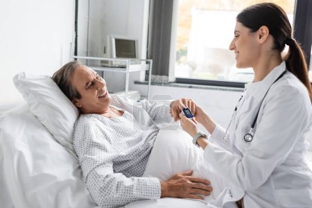 Positive doctor wearing pulse oximeter on finger of senior patient in hospital ward 