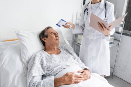 Doctor with paper folder holding pyrometer near senior patient in hospital 