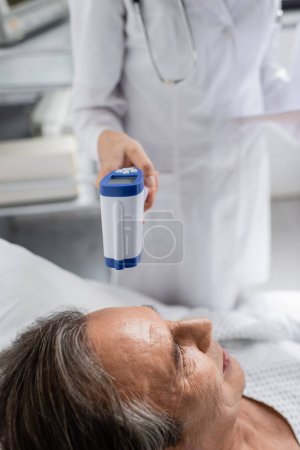 Verschwommener Arzt hält Pyrometer in der Nähe eines älteren Patienten auf Krankenhausstation 