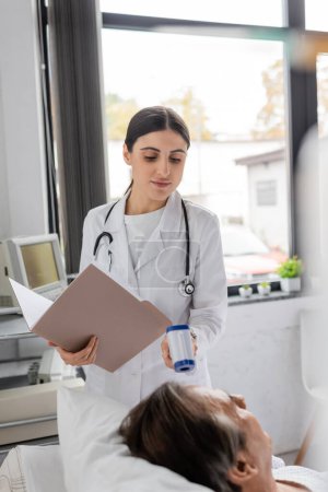 brunette doctor with paper folder checking temperature of senior patient in hospital ward 