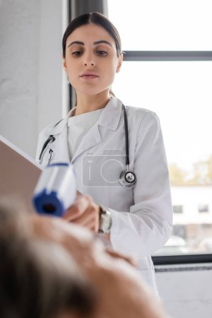 Doctor with paper folder holding pyrometer near blurred senior patient in clinic 
