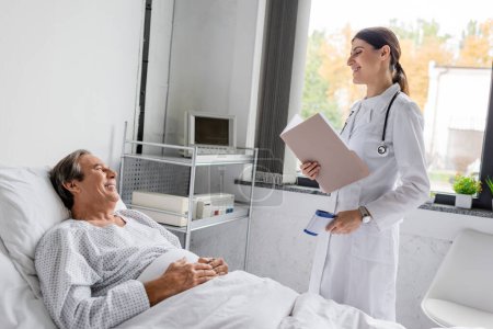 Positive elderly patient looking at doctor with paper folder and pyrometer in hospital ward 