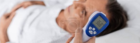 Doctor holding pyrometer near blurred patient on hospital bed, banner 