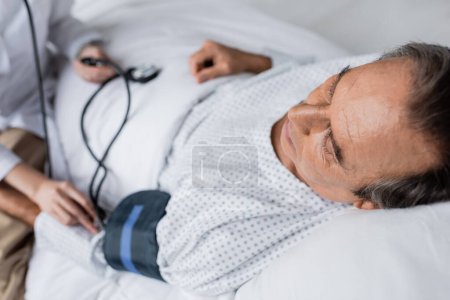 Kranker Patient liegt mit Tonometer neben verschwommenem Arzt in Klinik 