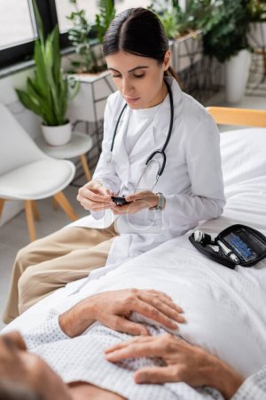 Doctor holding glucometer near senior patient on bed in clinic 