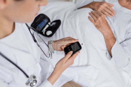 Ausgeschnittene Ansicht eines Arztes mit Glukometer in der Nähe kranker Seniorin im Krankenhaus 