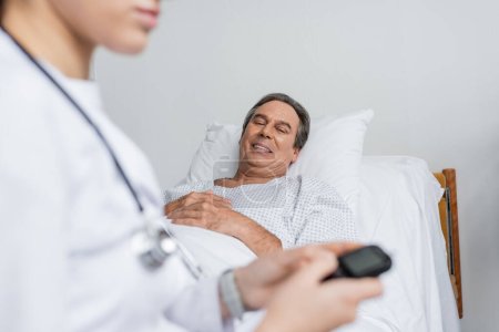 Lächelnder älterer Patient blickt verschwommenen Arzt mit Glukometer auf Krankenhausstation an 