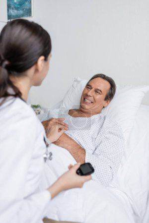 Smiling senior patient looking at blurred doctor with glucometer in clinic 