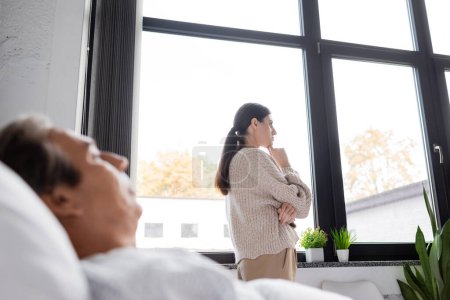 Téléchargez les photos : Side view of upset woman standing near blurred sick dad in hospital ward - en image libre de droit