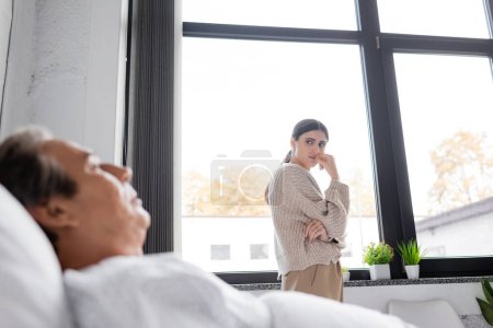 Traurige Frau sieht verschwommenen Vater in Klinik auf Bett liegen 