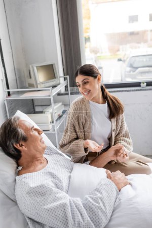 Positive woman holding hand of father in patient gown and talking in hospital ward 