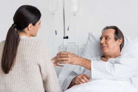 Blurred daughter giving glass of water to senior dad on hospital bed 