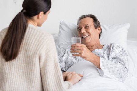 Smiling senior patient holding glass of water near blurred daughter in clinic 