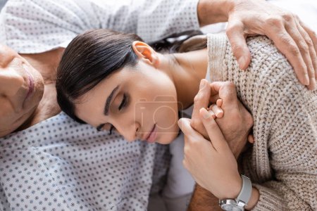 Top view of upset woman holding hand of sick father in hospital 